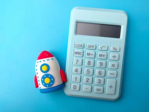 A toy rocket sits alongside a light blue electronic calculator on a blue su.. Stock Photos