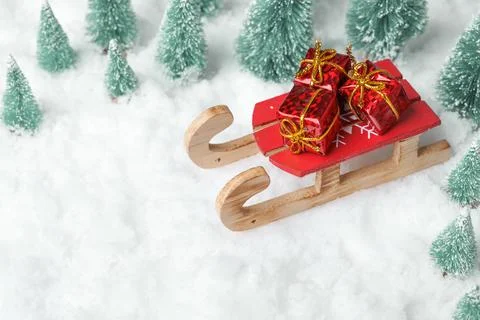Toy sleigh with three gift boxes on snow surrounded by miniature Christmas trees Stock Photos