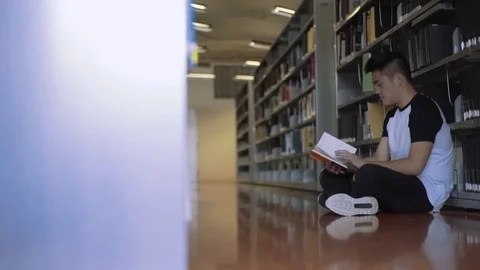 Tracking shot of boy sitting on the floor in library reading book Stock Footage