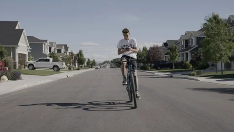 Tracking shot of distracted boy riding bicycle on suburban street while texting Stock Footage 117352629