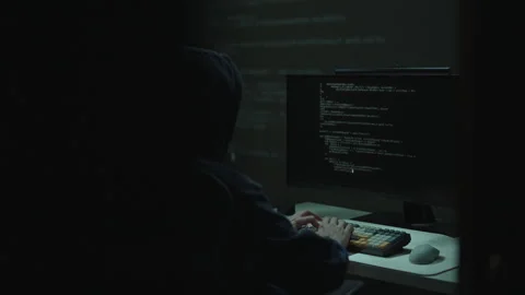 Tracking shot of hooded individual typing on keyboard, lines of code visible on Stock Footage 325711490