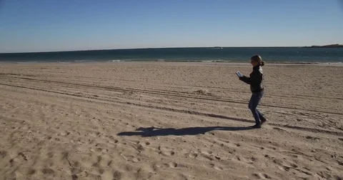 Tracking shot of lady walking on beach in Autumn with her tablet Stock Footage 71145698