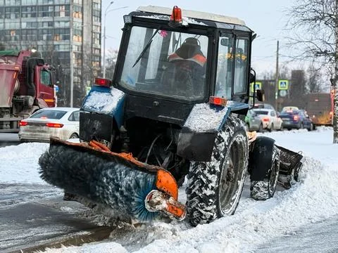 A tractor with a brush clears the road from snow. Streets in snow. Stock Photos