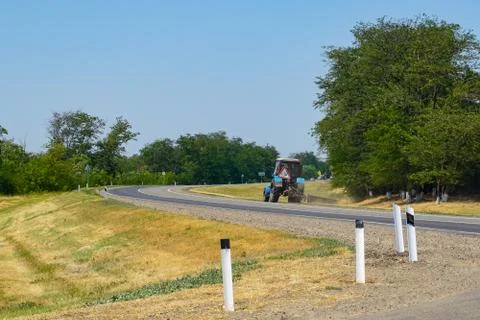 A tractor mows grass on the side of a highway near a field planted with wheat Stock Photos