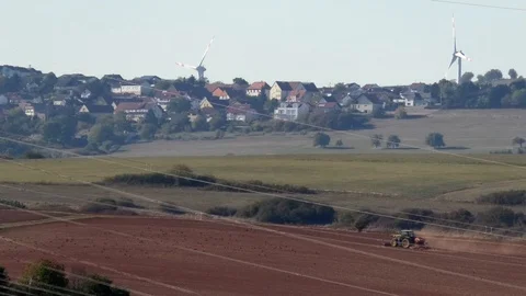 Tractor plowing field in front of rural German village Siegelbach, wind turbines Stock Footage 96006827