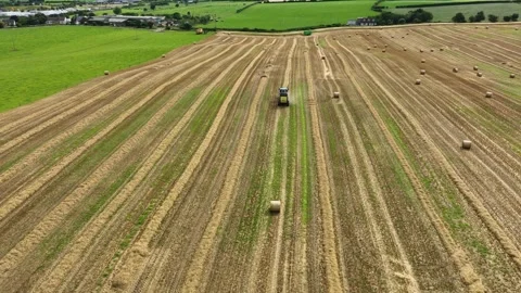 Tractor Pulling Round Baler Through Farmland With Village and Fields in 库存影片 341471590