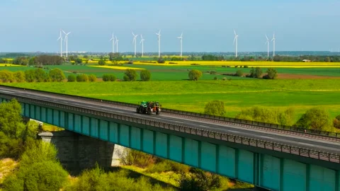 Tractor with sprayer driving over bridge with farmland and wind turbines in Stock Footage 311319816