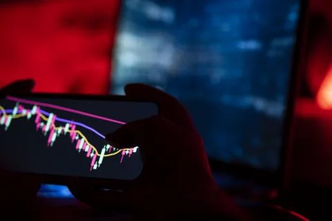 Trader holding a phone with a candlestick chart in a dark red-lit room Stock Photos