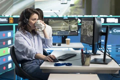 Traffic operator wearing headset drinks coffee while looking at camera footage Stock Photos