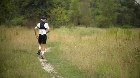 Trail runner wearing hydration backpack jogging down grassy path through meadow Stock Footage 313951345