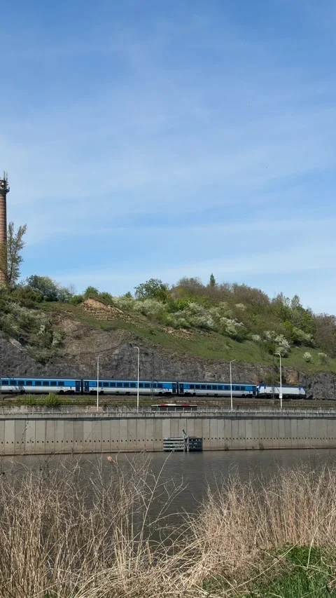 Train passing along riverside railway track, moving beside green hillside with Stock Footage 333699831
