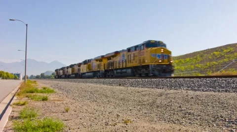 TRAIN PASSING BY ON RAILROAD NEAR OPEN FIELD ON A NICE SUNNY DAY WITH MOUNTAINS Stock Footage