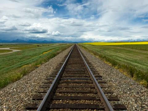 Train tracks between farm fields under a cloudy sky with Canadian Rockies in the Stock Photos