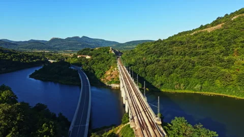 Train tracks run along a river with green hills in the background during a clear Stock Footage 342334897