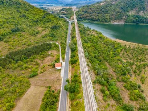 Train tracks run beside a road near a lake surrounded by hills and trees in.. 스톡 사진