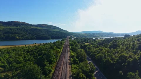 Train tracks run through green trees and hills beside a river with a blue sky Stock Footage 340765039