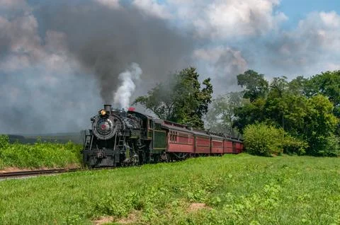 Train Travels Through Green Fields on a Sunny Day With Smoke Rising in the Sk Foto stock