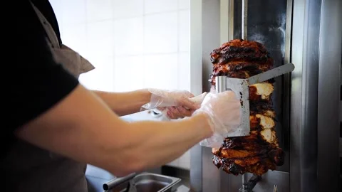 Trainee cook cuts grilled chicken meat from rotating spit and prepares Stock Footage 281248051