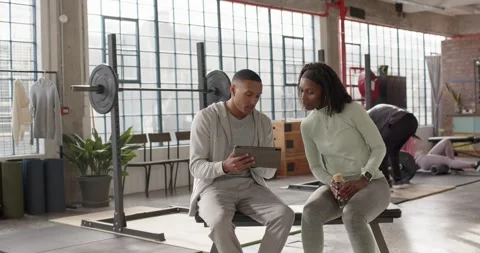 Trainer in gymwear opening tablet on bench, tapping, showing plan to female Stock Footage 337255251