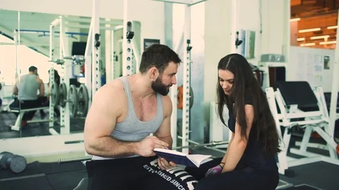 Trainer helps young strong brunette woman write training program in fitness club Stock Footage 72679280