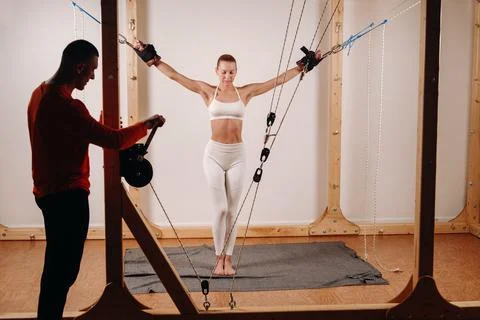 The trainer pulls the ropes stretching a Girl in a sports uniform on a simulator Stock Photos