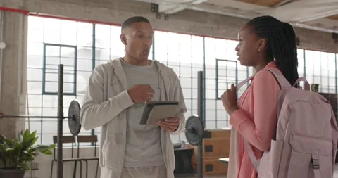 Trainer tapping tablet, gesturing, explaining gym plan to African American woman Stock Footage 337286707