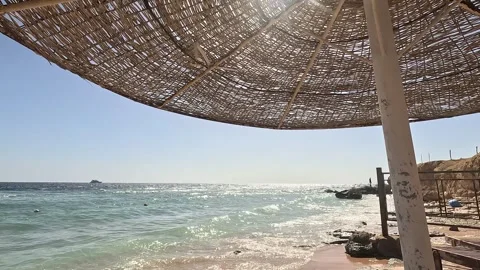 A Tranquil Beach Scene Under a Rustic Straw Umbrella Overlooking Gentle Waves Видео 330100286