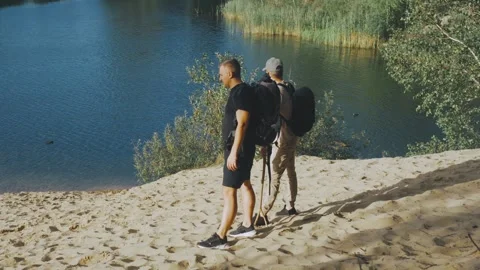 Tranquil pair exploring lakeshore with backpacks, Relaxed summer hiking along Stock Footage 323492584