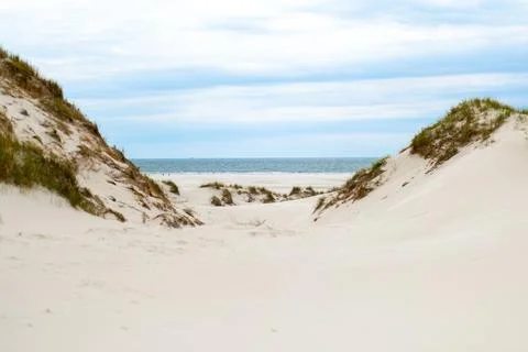A tranquil pathway through sand dunes leads to a serene beach under a cloudy sky Stock Photos