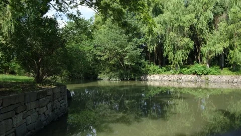 A tranquil pond with a stone wall along riverbank in a landscaped peaceful park Stock Footage 289625334