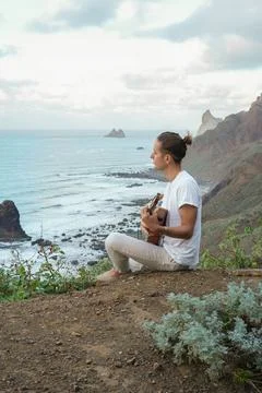 Tranquil Reflection: Young man Playing Ukulele on Cliff Edge Overlooking Ocean a Fotos de archivo