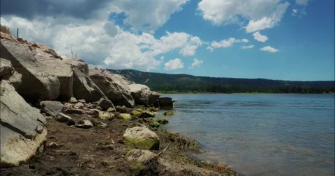 Tranquil River Flow by Rocky Shore Under Blue Sky 動画素材 324870245