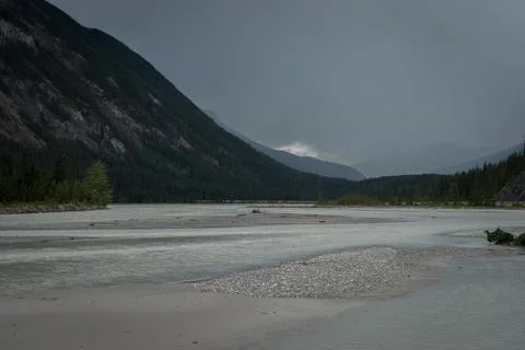 A tranquil river flows through a mountain valley under a cloudy sky. Stock Photos