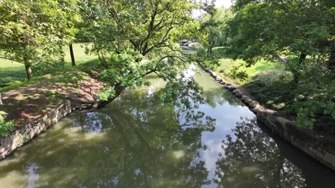 A tranquil stream winding through a lush green park with stone embankments Stock Footage 289622694