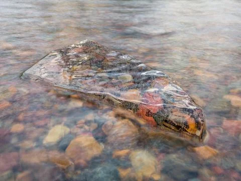 Translucent angular clean piece of ice in running water with colorful stones Stock Photos