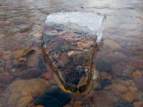 Translucent angular clean piece of ice in running water with colorful stones Stock Photos