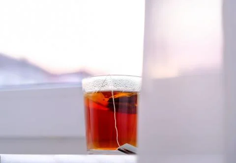 A transparent glass mug of black tea on white windowsill on sky background. M Stock Photos