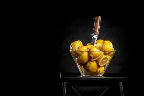 Transparent glass vase with yellow lemons and stuck knife on isolated black b Stock Photos