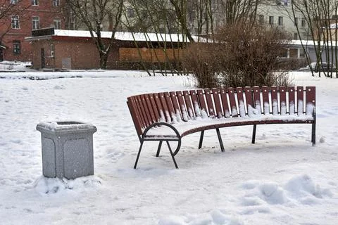 Trash can next to a bench covered with snow in a city park Stock Photos