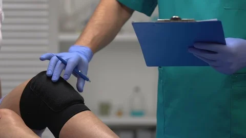 Traumatologist checking patients leg in knee brace, writing examination result Stock-Footage 117381233