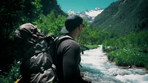 The traveler with a large backpack is standing by the mountain river and looks Stock Footage 138690348