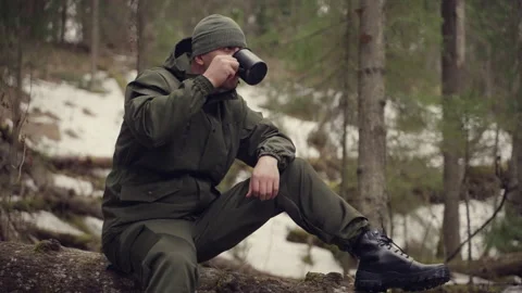 Traveler in a pine forest drinks coffee from a metal mug while sitting on a log Stockbeeldmateriaal 187101745