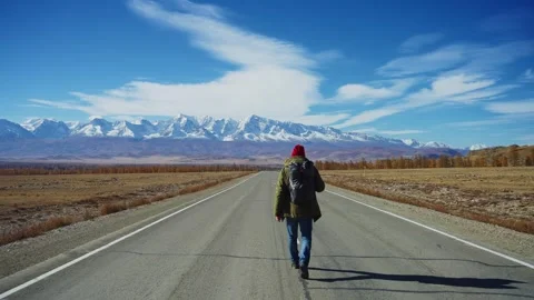Traveler in red hat strides forward on empty highway under vast open sky Stock Footage 342736160