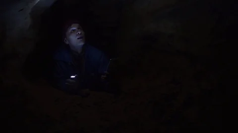Traveler using smartphone laying on ground in tight hole in cave with flashlight Stock Footage 80062760