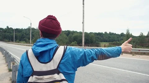 Traveller dressed in jacket and knitted hat with backpack hitchhiking on road Stock Footage 80054622