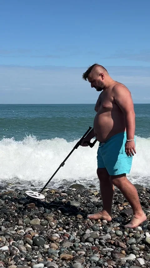Treasure hunter with big belly and metal detector walks on pebble sea beach Stock Footage 331172721