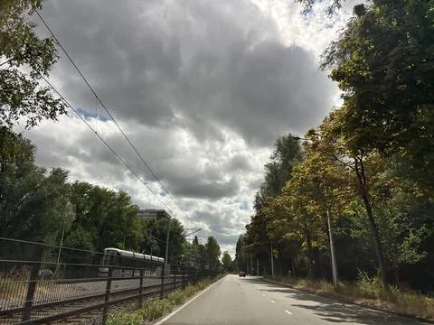 Tree lined road beside tram tracks under dramatic cloudy sky, with empty lanes Foto stock