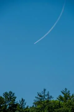Trees below a plane in the blue sky with a contrail Stock Photos