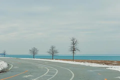 Trees line a path by the lake in winter with snow on the ground and water i.. Stock Photos