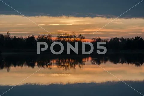 Trees on the shore of the lake and reflection of clouds after sunset Stock Photos